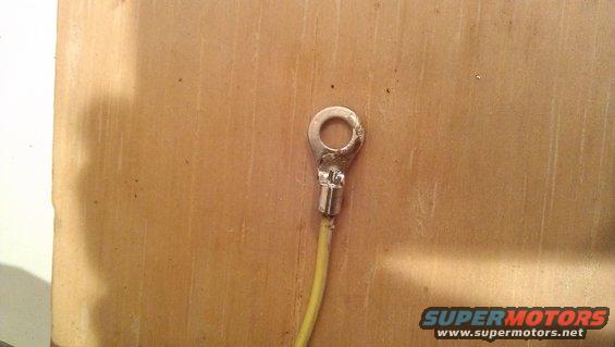 Electrical ring terminal crimped onto a yellow wire, against a wood-grain background