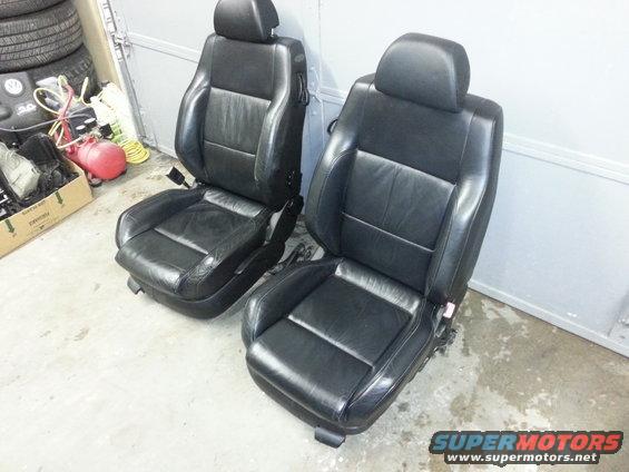 Two black leather car seats with headrests sit on a concrete floor. A box of parts and tires are in the background