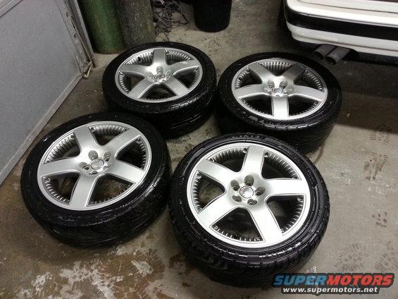 Four silver alloy wheels with black tires stacked in a garage. A white car's rear bumper and exhaust pipes are visible to the right
