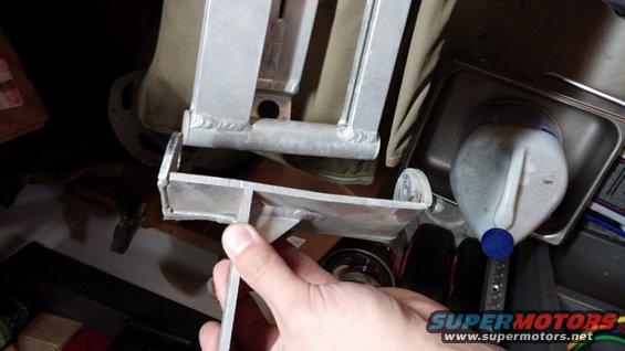 Hand holding a metal bracket with visible welds, next to a plastic jug in a metal container