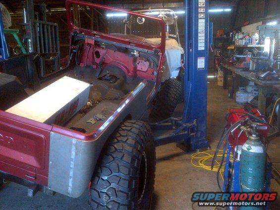 Red Jeep CJ-series body on a lift, rear view, showing disassembled interior and large off-road tires