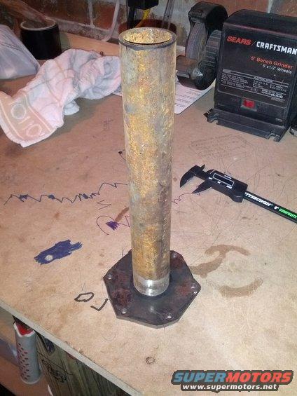 Rusted metal pipe mounted on a hexagonal metal plate, with a digital caliper and a bench grinder in the background