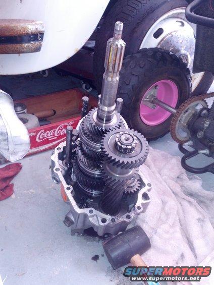 Transmission gears laid out on a white cloth next to a pink-rimmed wheel and a Coca-Cola crate