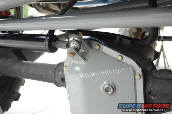 Close-up of a steering stabilizer and gray differential cover on a vehicle axle. Supermotors.net logo visible