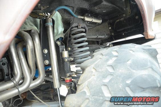 Close-up of off-road vehicle's front suspension with large coil spring, shock absorber, and exhaust headers. Large tire tread visible