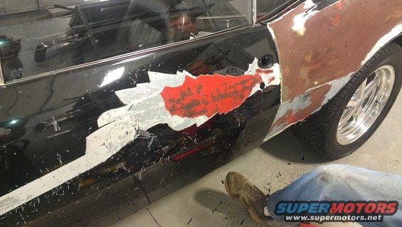 Car door with peeling black paint and red primer showing, fender is rusty and unpainted