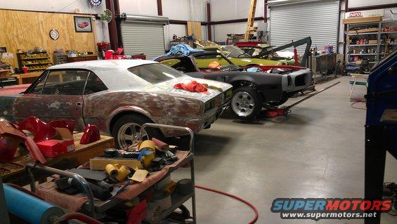 Three cars in various states of repair in a workshop. One car is a 1970s coupe, another is a black coupe with its front end disassembled, and a yellow coupe is in the background