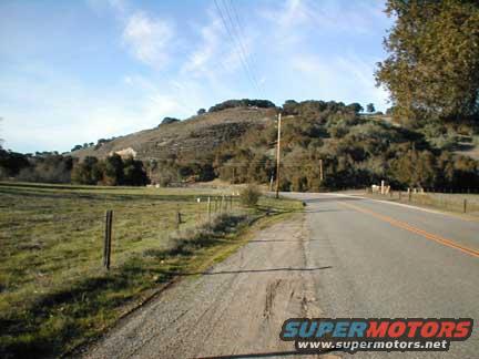 road curves through a grassy field toward a tree-covered hill under a blue sky