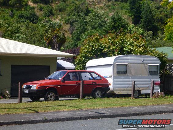 Red hatchback car parked next to a house, towing a silver trailer