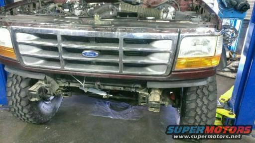 Front of a maroon Ford truck with hood open, showing grille and headlights. Tires and suspension visible