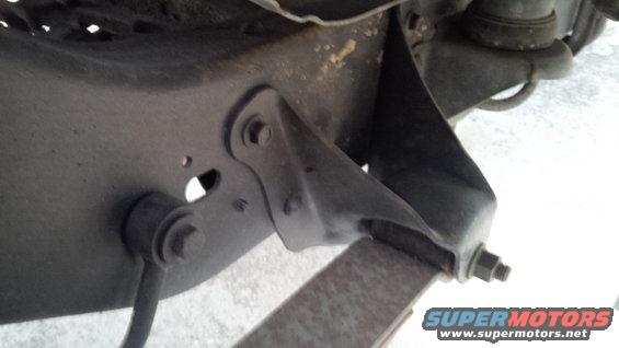Close-up of vehicle suspension parts, including a bracket and linkage, with a Supermotors.net watermark