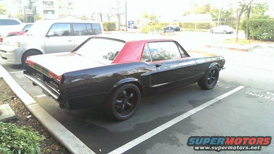 Black classic coupe with red roof and black wheels parked in a lot. Another white SUV is parked nearby