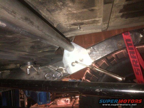 Underbody view of a vehicle showing custom suspension links and frame modifications. A red strap is visible