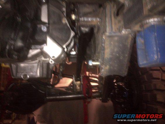 Underside view of a vehicle's drivetrain and suspension components. A blue container is visible on the right