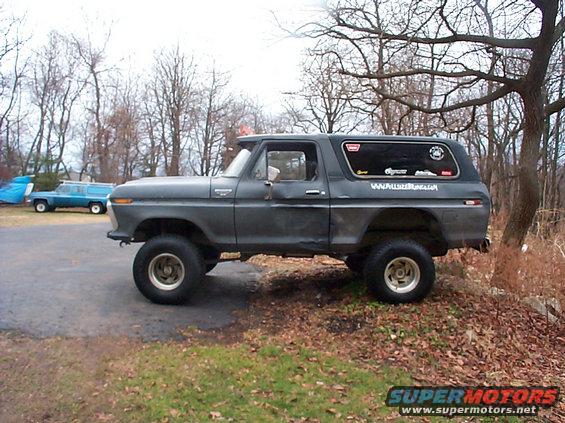 Gray 1978 Ford Bronco parked on a dirt incline, with a blue station wagon in the background