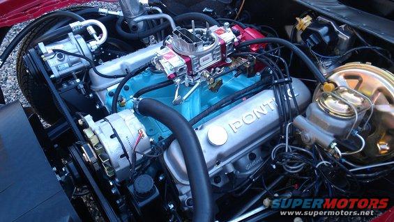 Close-up of a Pontiac V8 engine with blue block and silver valve covers. The 'Pontiac' logo is visible on the valve cover