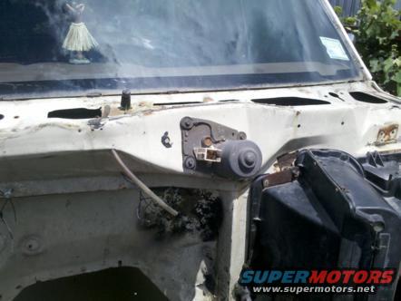 front cowl area of a vehicle, showing windshield wiper motor and surrounding parts. A hula dancer figurine is on the dashboard
