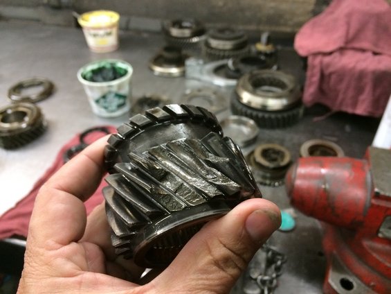 Hand holding a transmission gear with damaged teeth. Other gears and tools are visible on a workbench
