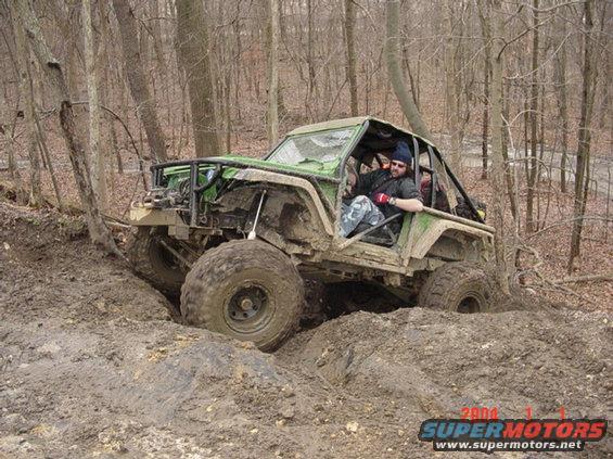 Modified off-road vehicle with large tires stuck in thick mud on a wooded trail