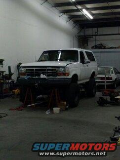 White Ford Bronco on jack stands, front end raised, in a workshop with another car in the background