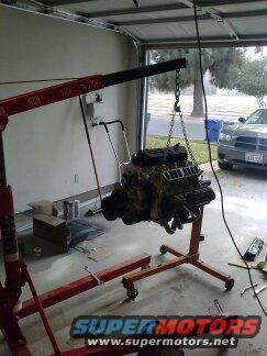 Engine hanging from an engine hoist in a garage. Another car is visible outside