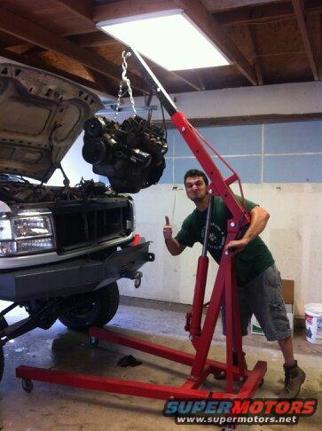 Gray truck with hood open, engine suspended by red engine hoist. Man gives thumbs up