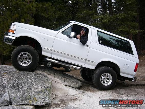White 1992 Ford Bronco with aftermarket wheels and side steps climbing over large rocks