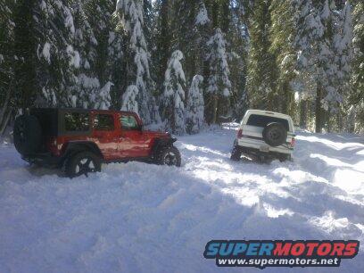 Red Jeep Wrangler and white SUV stuck in snow, surrounded by snow-covered trees