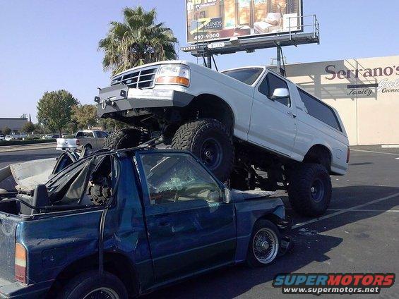 1995 Ford Bronco lifted high on top of a crushed blue pickup truck