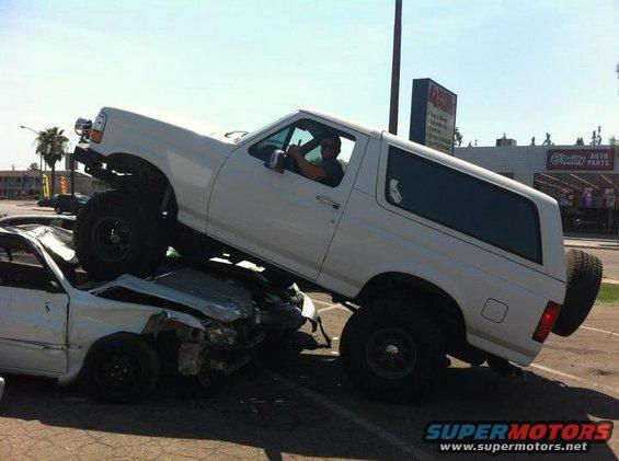 White Ford Bronco on top of a wrecked white car, driver giving thumbs up