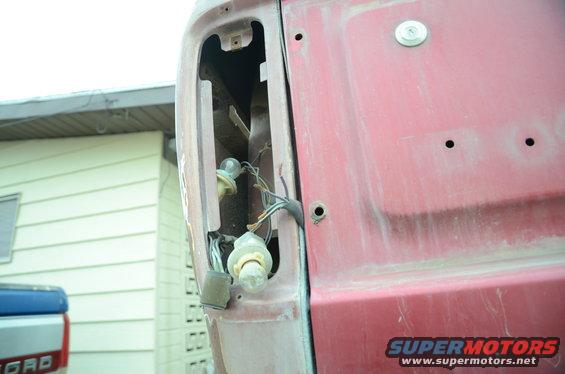 Red tailgate section of a 1979 Ford Bronco with exposed wiring and light sockets, showing a blue truck bed in the background