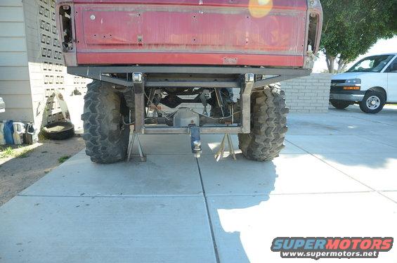 red pickup truck rear end on jack stands, showing custom hitch and large tires. White van in background