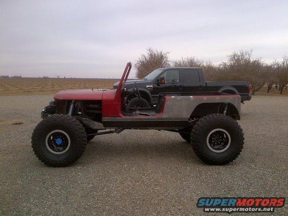 Red Jeep CJ-style body with exposed frame, large tires, and a black pickup truck behind it