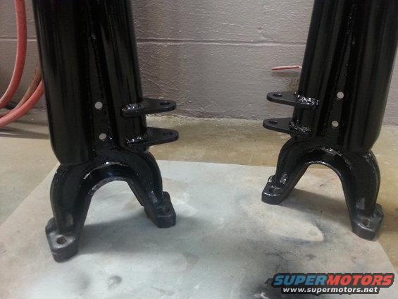 Two black metal automotive parts with mounting flanges sit on a concrete floor