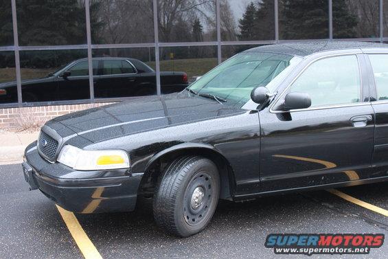 Black sedan parked next to a building with a reflection of another black sedan in the windows. Yellow lines on wet asphalt