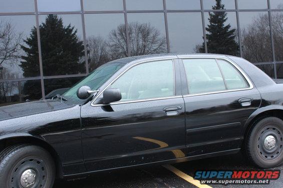 Side view of a black sedan parked in front of a building with tinted windows. The car has steel wheels and a chrome trim around the windows