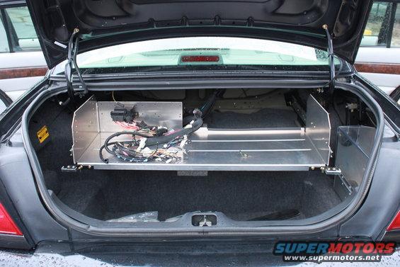 Black car trunk interior with aluminum shelving and exposed wiring. A yellow sticker is visible on the left wall