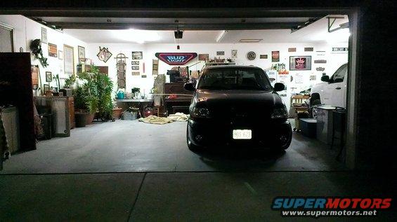 Black pickup truck parked in a garage with a white truck behind it. License plate is visible