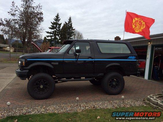 Black Ford Bronco with blue stripe, lifted with large tires. Fire Dept flag flying