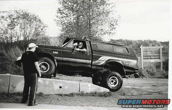 Dark Ford F-150 pickup truck with a camper shell, angled on a concrete barrier. A person is in the driver's seat