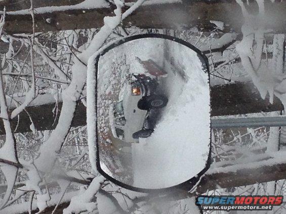 Reflection in a snow-covered truck side mirror shows a dark vehicle with an orange turn signal, partially obscured by snow