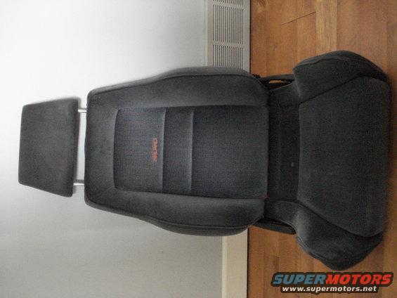 Dark gray fabric car seat with orange RECARO logo, detached headrest, on wooden floor