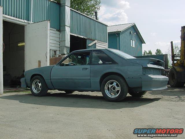Gray 1990 Ford Mustang coupe with aftermarket wheels, primer finish, parked outside a garage