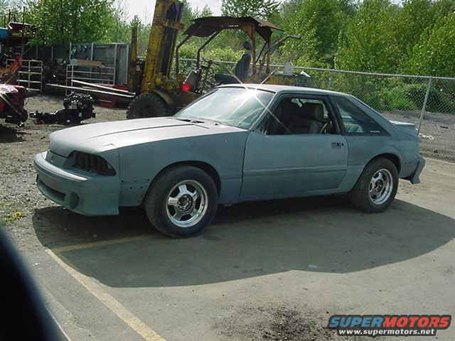 Gray Ford Mustang coupe, primer coat, parked outside with a forklift in background