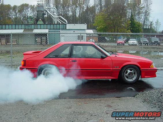 Red coupe with a spoiler doing a burnout, emitting white smoke from the rear tires