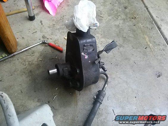 power steering pump sitting on concrete floor, with a rag stuffed in top opening. Tools and a jack stand are nearby