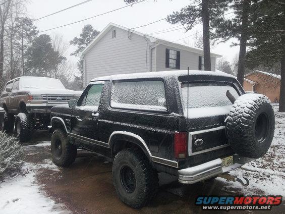 Two Ford Broncos parked in snow, one black with spare tire on back, another brown on trailer