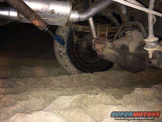 Rear axle, driveshaft, and exhaust of a vehicle in sand. The driveshaft has a blue protective cap
