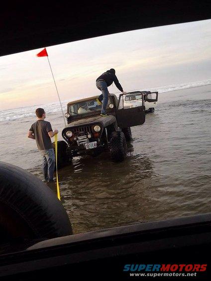 Two Jeeps partially submerged in water on a beach, one with a person climbing on it and another with the door open