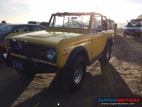 Yellow first-generation Ford Bronco with aftermarket bumper and lights parked on sand. Other vehicles visible in background